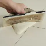 Main tenant utilise une spatule en bois pour faire des joints de carrelage, étalant le mortier sur les carreaux clairs.