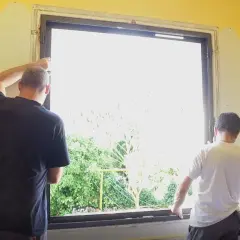 Deux hommes installent une nouvelle fenêtre. La lumière du jour filtre à travers le vitrage, révélant un jardin verdoyant. La pièce est encore en travaux, mais bientôt, cette fenêtre dormante s'ouvrira sur le monde.