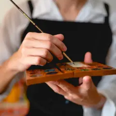 Main tenant une palette en bois remplie de couleurs, un artiste utilise un pinceau, un des outils pour peindre, pour mélanger des pigments.