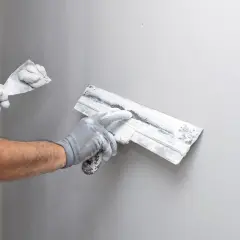 Mains gantées appliquant de l'enduit sur un mur gris, étape clé de la construction murale.