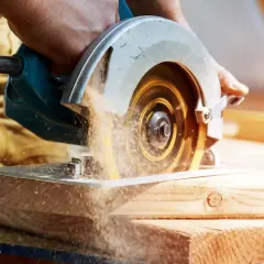 Main d'un bricoleur utilisant une scie circulaire pour couper du bois. La poussière de bois vole autour de la lame. Apprenez comment affûter une lame de scie circulaire pour des coupes nettes.