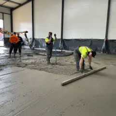 Des ouvriers coulent et lissent du béton frais sur un sol, avec une pompe à béton en arrière-plan.