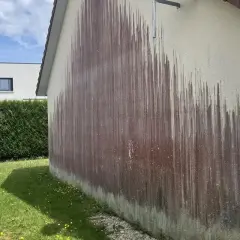 Le mur extérieur d'une maison présente une importante moisissure rouge, formant des traînées sombres sur le crépi clair.