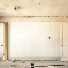 Intérieur d'une pièce abandonnée, papier peint décoloré, plafond avec des taches d'humidité et de la peinture écaillée, sol jonché de débris.