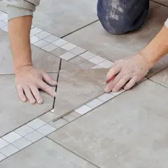 Mains expertes en pose carrelage, ajustant une pièce avec précision. Un coupe-carreau orange est visible.