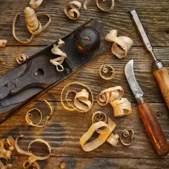 Un rabot, un ciseau et un couteau à bois, outils pour travailler le bois à la main, entourés de copeaux sur un établi rustique.