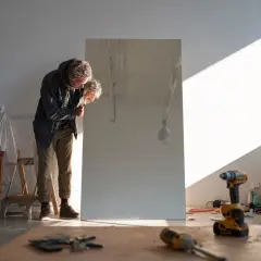 Un homme se penche pour accrocher un miroir au mur dans son atelier. Des outils sont posés au sol.