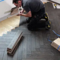 Pose de parquet en pose bâton rompu. Un artisan applique de la colle sur le sol avant de poser les lames de bois.