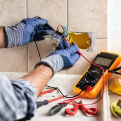 Un électricien vérifie le **fonctionnement d'une gâche électrique** avec un multimètre. Outils et casque de sécurité à proximité.