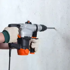 Une personne en salopette bleue, portant des gants, utilise un perforateur pour percer un mur en béton.