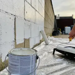 Un rouleau blanc est utilisé pour peindre un mur en parpaing avec de la peinture blanche. Une boîte de peinture est visible au premier plan.