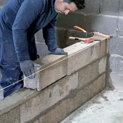Un maçon pose une brique sur un mur en agglo, guidé par un fil à plomb. Outils de maçonnerie à proximité.