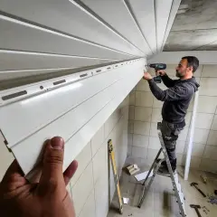 Un homme sur une échelle réalise la pose de lambris PVC au plafond. Des outils sont visibles au sol.