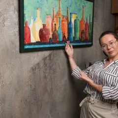 Une femme montre comment accrocher un cadre coloré de bouteilles sur un mur texturé.