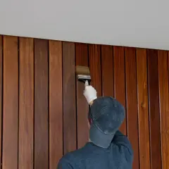 Une personne porte un gant blanc et un casquette pour peindre du lambris en bois avec un pinceau.