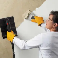 Une femme, portant des gants jaunes, utilise un appareil pour décoller du papier peint ancien. Elle gratte la surface avec un outil.
