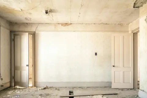 Intérieur d'une pièce abandonnée, papier peint décoloré, plafond avec des taches d'humidité et de la peinture écaillée, sol jonché de débris.