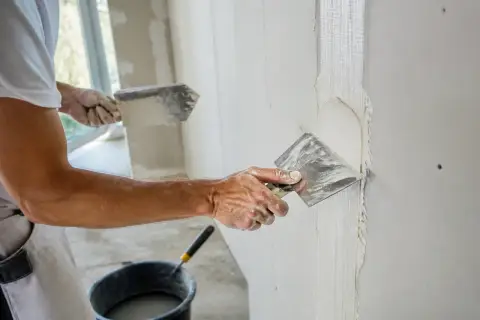 Un ouvrier applique de l'enduit sur une bande de placo. La question "faut-il humidifier les bandes placo" est au cœur de cette étape de finition.
