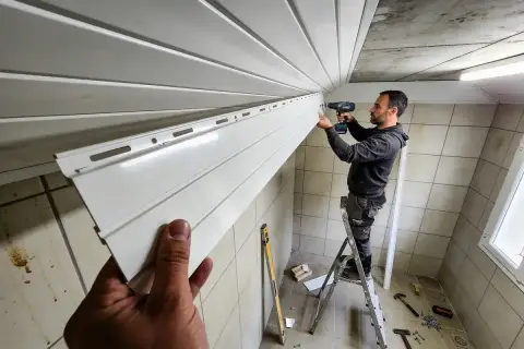 Un homme sur une échelle réalise la pose de lambris PVC au plafond. Des outils sont visibles au sol.