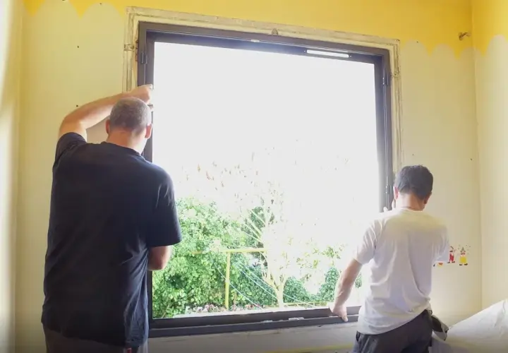 Deux hommes installent une nouvelle fenêtre. La lumière du jour filtre à travers le vitrage, révélant un jardin verdoyant. La pièce est encore en travaux, mais bientôt, cette fenêtre dormante s'ouvrira sur le monde.
