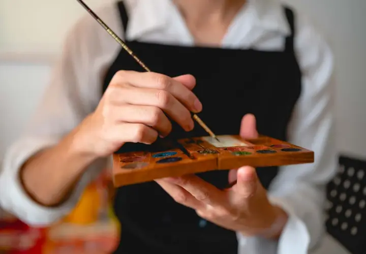 Main tenant une palette en bois remplie de couleurs, un artiste utilise un pinceau, un des outils pour peindre, pour mélanger des pigments.