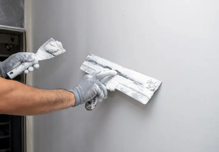 Mains gantées appliquant de l'enduit sur un mur gris, étape clé de la construction murale.