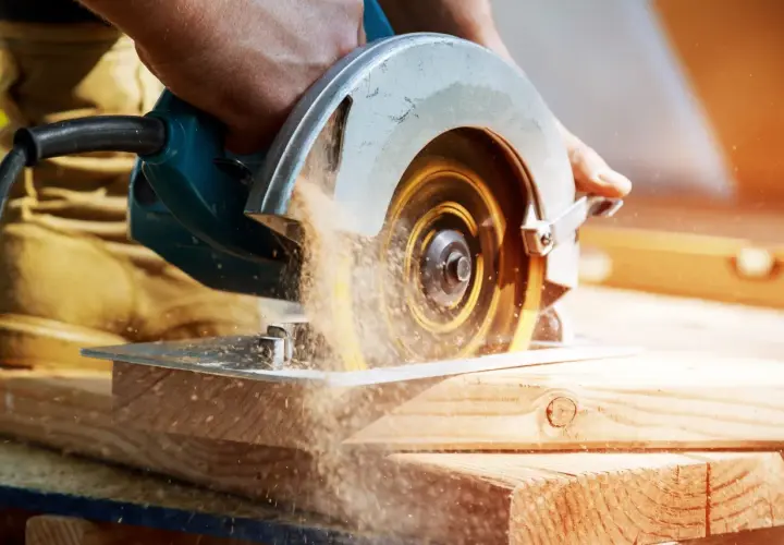 Main d'un bricoleur utilisant une scie circulaire pour couper du bois. La poussière de bois vole autour de la lame. Apprenez comment affûter une lame de scie circulaire pour des coupes nettes.