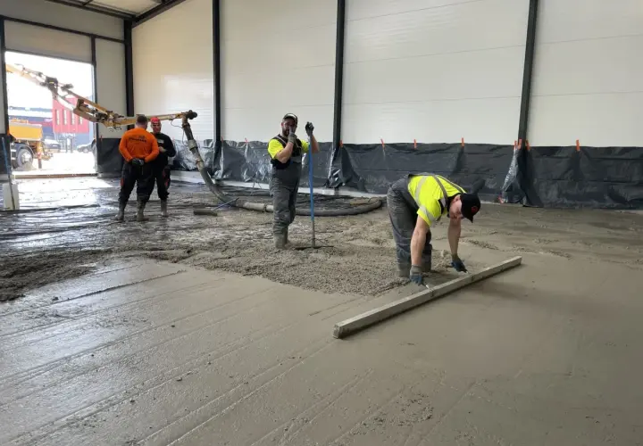 Des ouvriers coulent et lissent du béton frais sur un sol, avec une pompe à béton en arrière-plan.