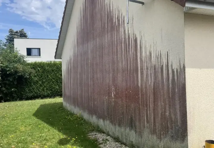 Le mur extérieur d'une maison présente une importante moisissure rouge, formant des traînées sombres sur le crépi clair.