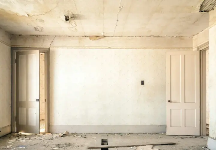 Intérieur d'une pièce abandonnée, papier peint décoloré, plafond avec des taches d'humidité et de la peinture écaillée, sol jonché de débris.