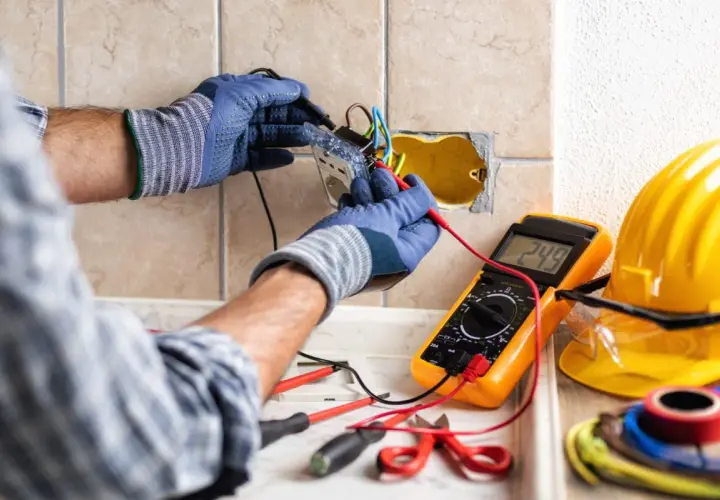 Un électricien vérifie le **fonctionnement d'une gâche électrique** avec un multimètre. Outils et casque de sécurité à proximité.