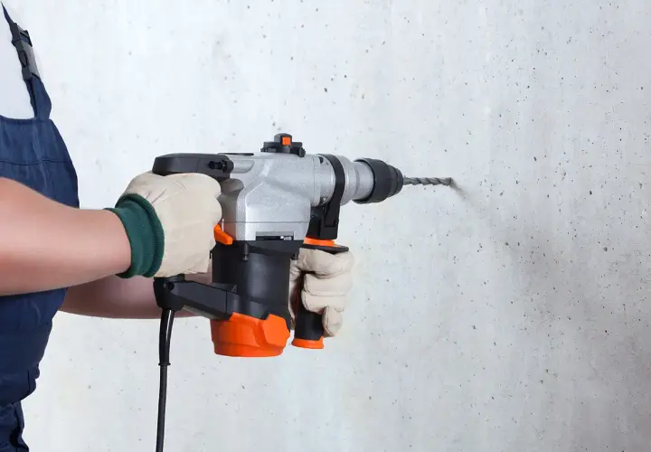 Une personne en salopette bleue, portant des gants, utilise un perforateur pour percer un mur en béton.