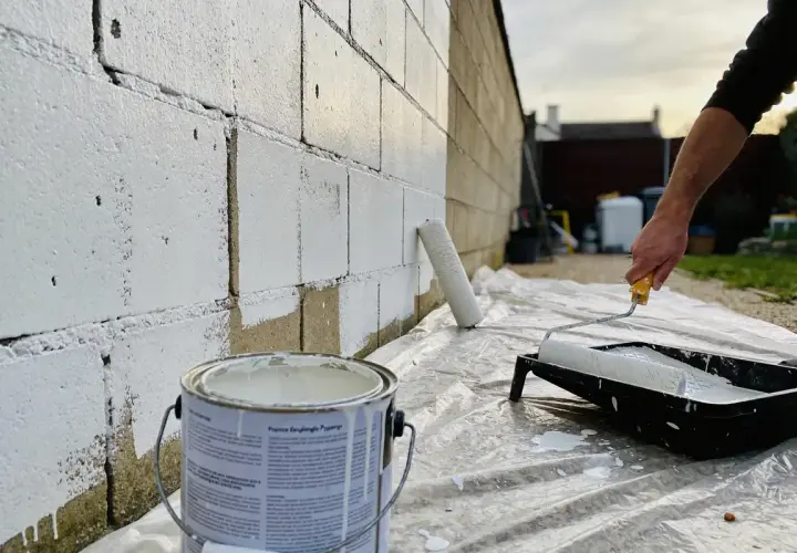 Un rouleau blanc est utilisé pour peindre un mur en parpaing avec de la peinture blanche. Une boîte de peinture est visible au premier plan.