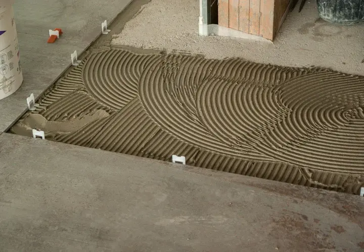 La chape est prête à recevoir les carreaux, avec un mortier étalé en ondulations parfaites. Des croisillons blancs maintiennent l'espacement.