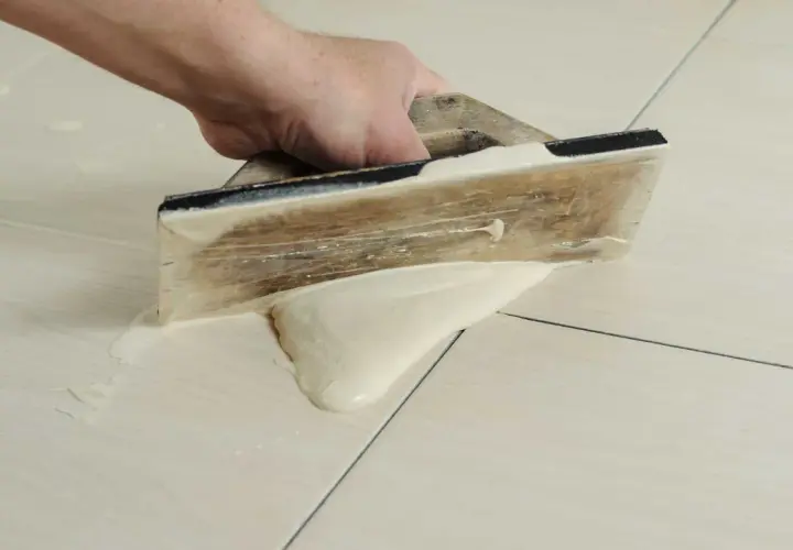 Main tenant utilise une spatule en bois pour faire des joints de carrelage, étalant le mortier sur les carreaux clairs.