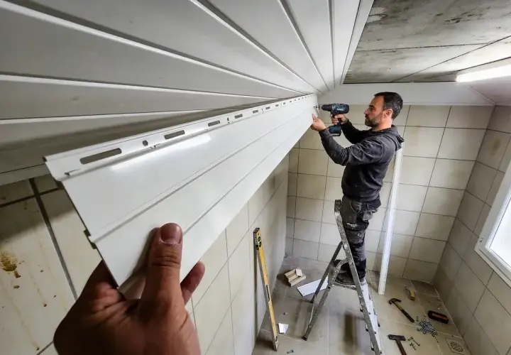 Un homme sur une échelle réalise la pose de lambris PVC au plafond. Des outils sont visibles au sol.