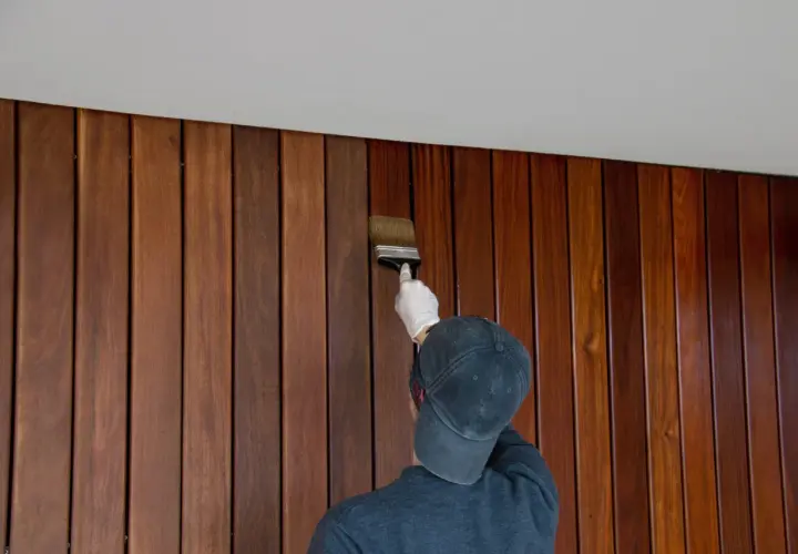 Une personne porte un gant blanc et un casquette pour peindre du lambris en bois avec un pinceau.