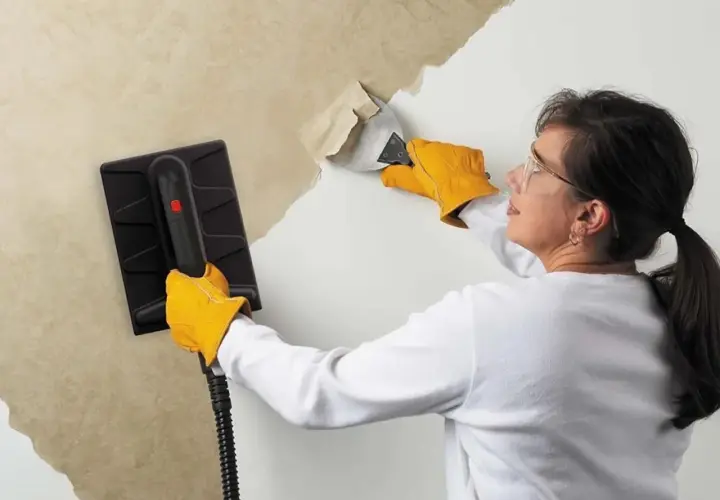 Une femme, portant des gants jaunes, utilise un appareil pour décoller du papier peint ancien. Elle gratte la surface avec un outil.