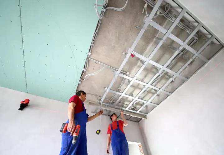 Deux ouvriers installent une structure métallique pour un schema montage placo plafond. Des fils électriques sont déjà en place.