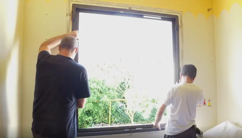 Deux hommes installent une nouvelle fenêtre. La lumière du jour filtre à travers le vitrage, révélant un jardin verdoyant. La pièce est encore en travaux, mais bientôt, cette fenêtre dormante s'ouvrira sur le monde.