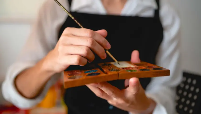 Main tenant une palette en bois remplie de couleurs, un artiste utilise un pinceau, un des outils pour peindre, pour mélanger des pigments.