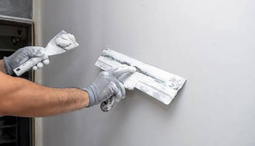 Mains gantées appliquant de l'enduit sur un mur gris, étape clé de la construction murale.