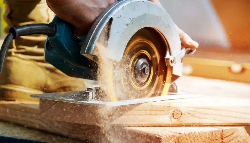 Main d'un bricoleur utilisant une scie circulaire pour couper du bois. La poussière de bois vole autour de la lame. Apprenez comment affûter une lame de scie circulaire pour des coupes nettes.