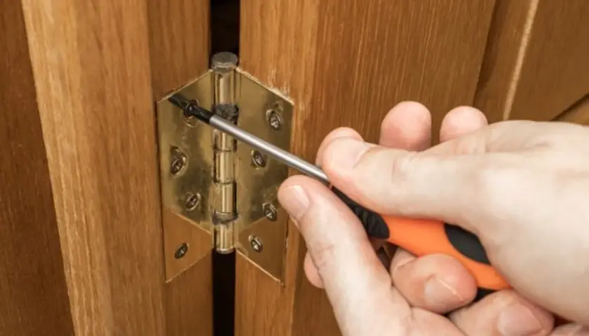 Main tenant une visseuse, une personne ajuste une charnière plate sur une porte en bois.