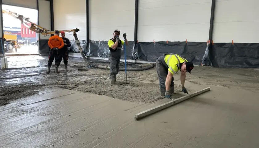 Des ouvriers coulent et lissent du béton frais sur un sol, avec une pompe à béton en arrière-plan.