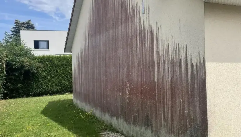Le mur extérieur d'une maison présente une importante moisissure rouge, formant des traînées sombres sur le crépi clair.