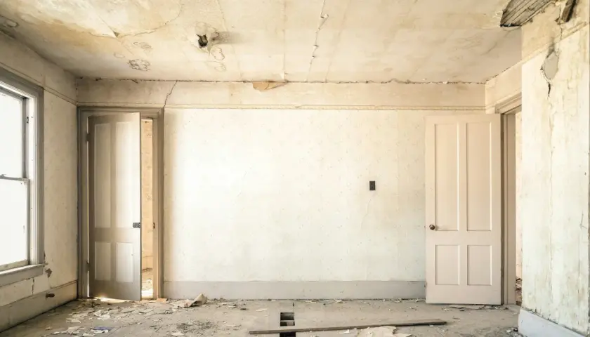 Intérieur d'une pièce abandonnée, papier peint décoloré, plafond avec des taches d'humidité et de la peinture écaillée, sol jonché de débris.
