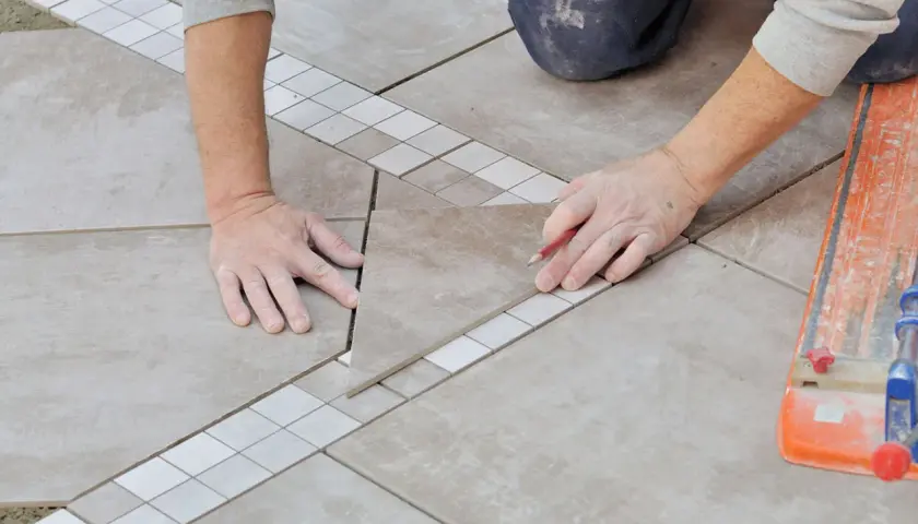 Mains expertes en pose carrelage, ajustant une pièce avec précision. Un coupe-carreau orange est visible.
