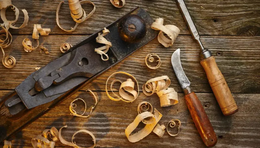 Un rabot, un ciseau et un couteau à bois, outils pour travailler le bois à la main, entourés de copeaux sur un établi rustique.