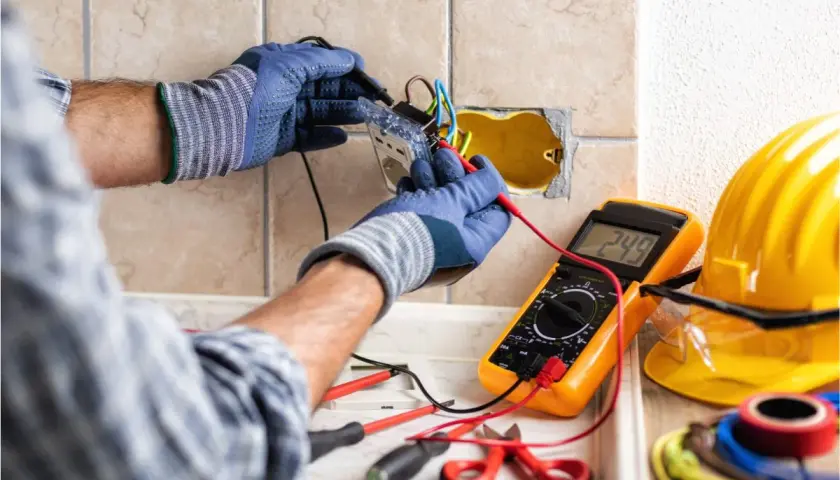 Un électricien vérifie le **fonctionnement d'une gâche électrique** avec un multimètre. Outils et casque de sécurité à proximité.